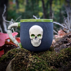 May include: A dark gray ceramic mug featuring a raised, off-white human skull relief on the front. The mug has a bright green rim and a handle sculpted to resemble a bone. The interior of the skull's eye sockets is glazed in a matching green.