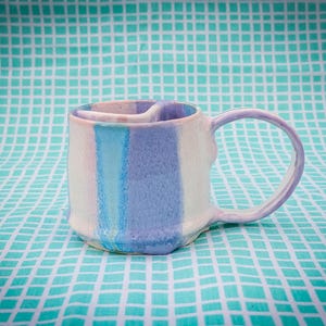 May include: A handmade ceramic mug with a unique design. The mug features a white base with vertical stripes of light blue and lavender. The handle and interior rim are lavender. The mug is sitting on a turquoise and white checkered surface.