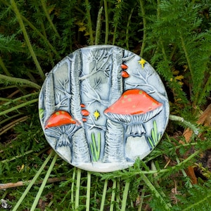 May include: A round ceramic piece featuring a forest scene with two large red-capped mushrooms with white spots. The background shows stylized trees and stars, with green grass accents. The piece is set against a backdrop of green foliage.