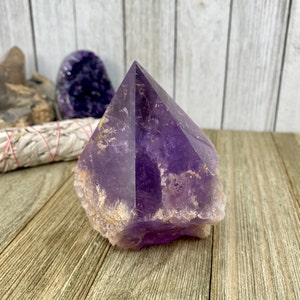 K&ouml;nnte beinhalten: Eine gro&szlig;e, violette Amethyst-Kristallspitze mit einer nat&uuml;rlichen, rauen Textur. Der Kristall ist klar und hat einen leichten goldenen Farbton. Er liegt auf einer Holzoberfl&auml;che.