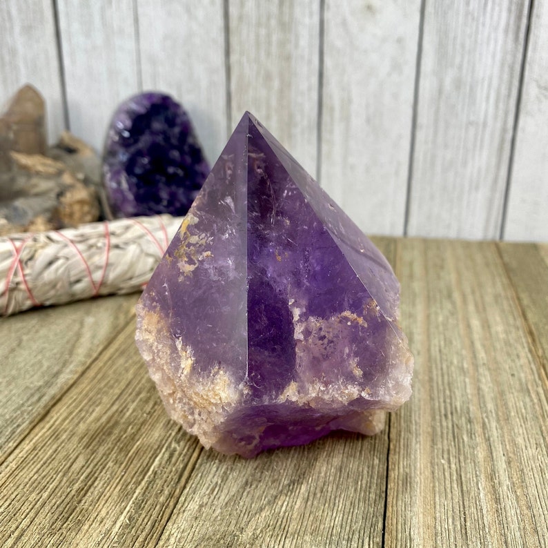 K&ouml;nnte beinhalten: Eine gro&szlig;e, violette Amethyst-Kristallspitze mit einer nat&uuml;rlichen, rauen Textur. Der Kristall ist klar und hat einen leichten goldenen Farbton. Er liegt auf einer Holzoberfl&auml;che.