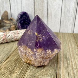 K&ouml;nnte beinhalten: Eine gro&szlig;e, violette Amethyst-Kristallspitze mit einer rauen, nat&uuml;rlichen Basis. Der Kristall ist klar und hat einen leichten goldenen Farbton.