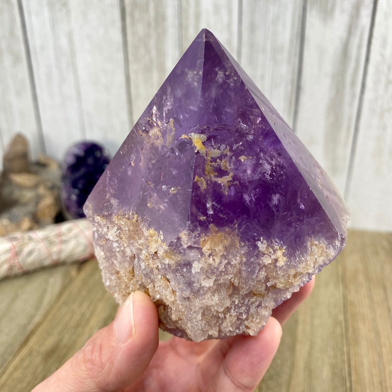 K&ouml;nnte beinhalten: Eine gro&szlig;e, violette Amethyst-Kristallspitze mit einer nat&uuml;rlichen, rauen Basis. Der Kristall ist klar und hat einen hellvioletten Farbton. Die Basis ist mit kleineren Kristallen bedeckt.