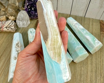 Peut inclure: Une tour en cristal poli de forme hexagonale présentant des bandes de couches minérales bleu clair, blanches et beige. La pierre possède une surface lisse et translucide avec des stries naturelles visibles et une pointe effilée.