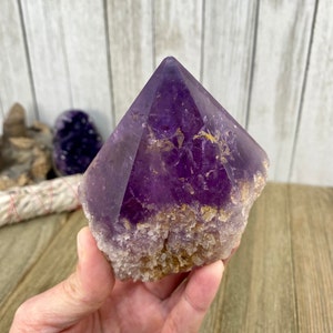 K&ouml;nnte beinhalten: Eine gro&szlig;e, violette Amethyst-Kristallspitze mit einer nat&uuml;rlichen, rauen Basis. Der Kristall ist klar und hat eine tiefviolette Farbe. Die Basis ist mit kleineren Kristallen bedeckt.