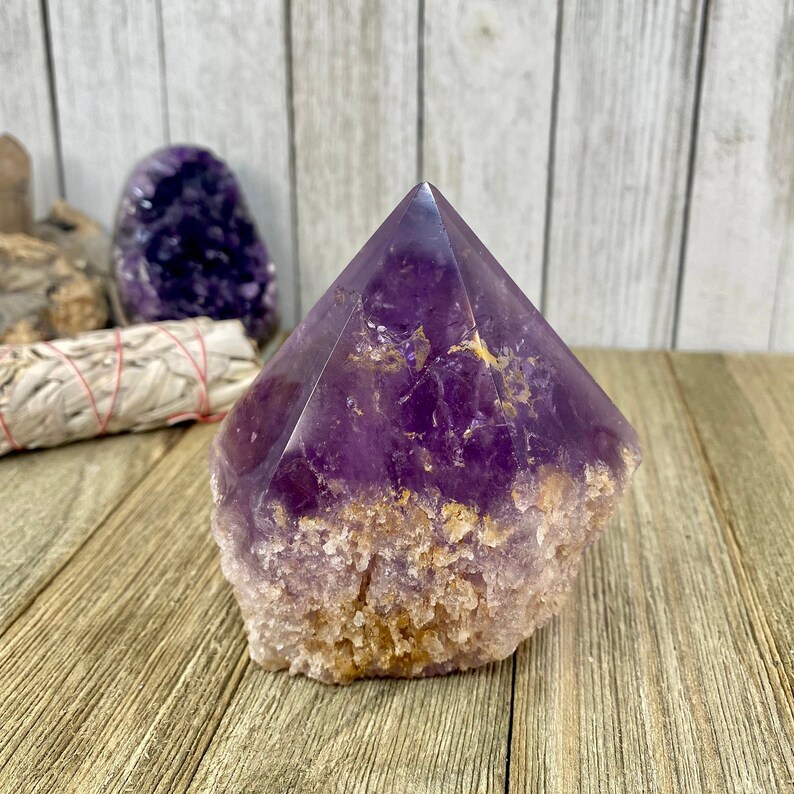 K&ouml;nnte beinhalten: Eine gro&szlig;e, violette Amethyst-Kristallspitze mit einer rauen, strukturierten Basis. Der Kristall ist klar und hat einen leichten goldenen Farbton.