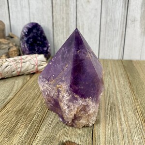 K&ouml;nnte beinhalten: Eine gro&szlig;e, violette Amethyst-Kristallspitze mit einer nat&uuml;rlichen, rauen Textur. Der Kristall ist pyramidenf&ouml;rmig und hat eine tiefe, satte violette Farbe.