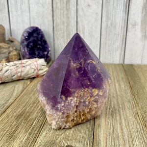 K&ouml;nnte beinhalten: Eine gro&szlig;e, violette Amethyst-Kristallspitze mit einer nat&uuml;rlichen, rauen Basis. Der Kristall ist klar und hat einen leichten goldenen Farbton.
