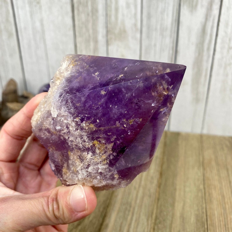 K&ouml;nnte beinhalten: Ein gro&szlig;er, violetter Amethystkristall mit einer spitzen Spitze und einer rauen, nat&uuml;rlichen Oberfl&auml;che. Der Kristall ist durchscheinend und hat eine tiefviolette Farbe.