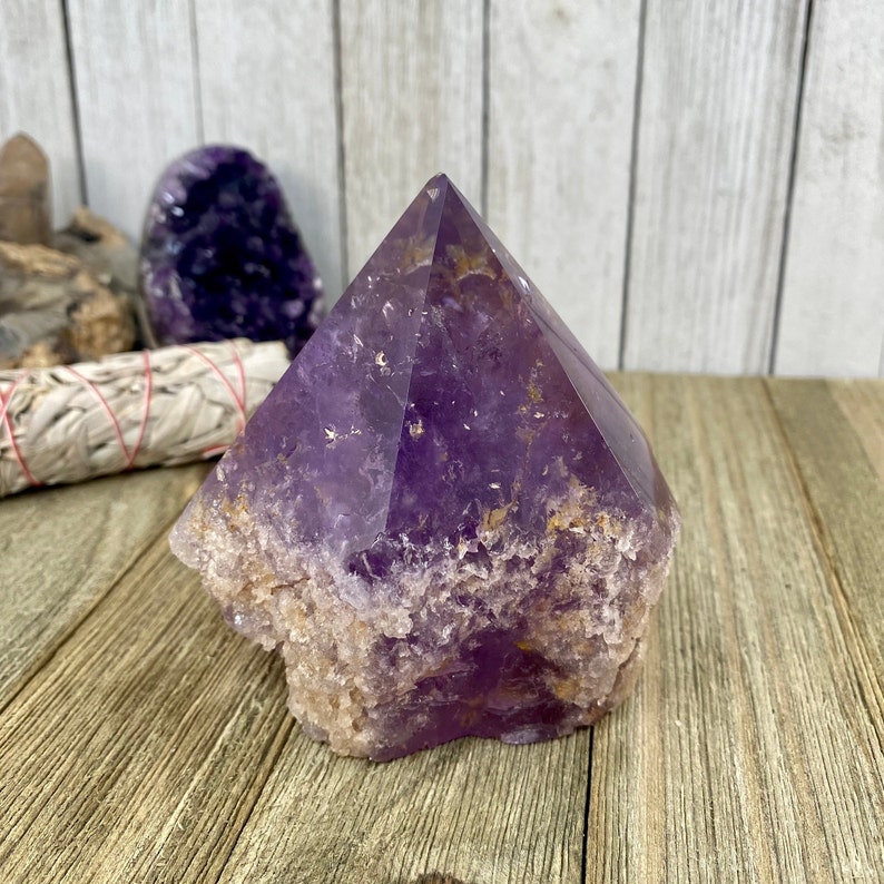 K&ouml;nnte beinhalten: Eine gro&szlig;e, violette Amethyst-Kristallspitze mit einer rauen, nat&uuml;rlichen Textur. Der Kristall ist pyramidenf&ouml;rmig und hat eine Spitze oben. Der Kristall liegt auf einer Holzoberfl&auml;che.