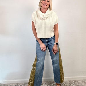 May include: A woman wearing a cream-colored, short-sleeved sweater with a cowl neck and blue jeans. The jeans have olive green panels on the sides with white text and decorative stitching. She is wearing brown sandals.