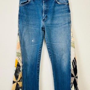 Op de afbeelding: Blauwe spijkerbroek met patchwork stoffen accenten aan de zijkanten. Het patchwork heeft een verscheidenheid aan kleuren en patronen, waaronder geel, zwart en cr&egrave;me. De jeans heeft een klassiek ontwerp met vijf zakken en een rechte pijp.