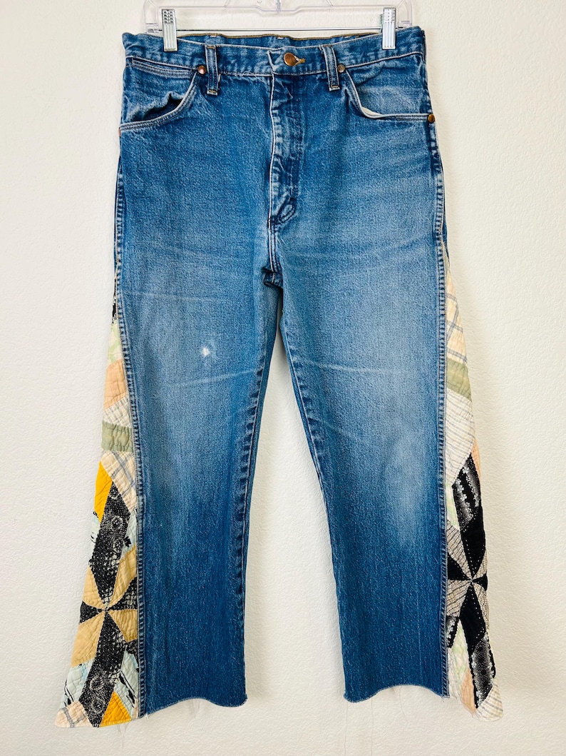 Op de afbeelding: Blauwe spijkerbroek met patchwork stoffen accenten aan de zijkanten. Het patchwork heeft een verscheidenheid aan kleuren en patronen, waaronder geel, zwart en cr&egrave;me. De jeans heeft een klassiek ontwerp met vijf zakken en een rechte pijp.
