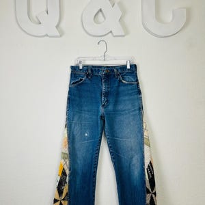 Op de afbeelding: Blauwe spijkerbroek met patchwork accenten aan de zijkanten. Het patchwork heeft een verscheidenheid aan kleuren en patronen, waaronder geel, zwart en cr&egrave;me. De jeans hangt aan een doorzichtige hanger tegen een witte achtergrond.