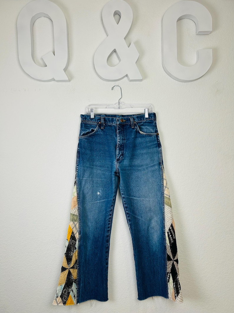 Op de afbeelding: Blauwe spijkerbroek met patchwork accenten aan de zijkanten. Het patchwork heeft een verscheidenheid aan kleuren en patronen, waaronder geel, zwart en cr&egrave;me. De jeans hangt aan een doorzichtige hanger tegen een witte achtergrond.