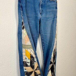 Op de afbeelding: Blauwe spijkerbroek met patchwork quilt details aan de zijkanten. Het patchwork heeft verschillende kleuren en patronen, waaronder beige, geel, zwart en roze. De jeans hebben een klassiek ontwerp met vijf zakken en een knoop- en ritssluiting.