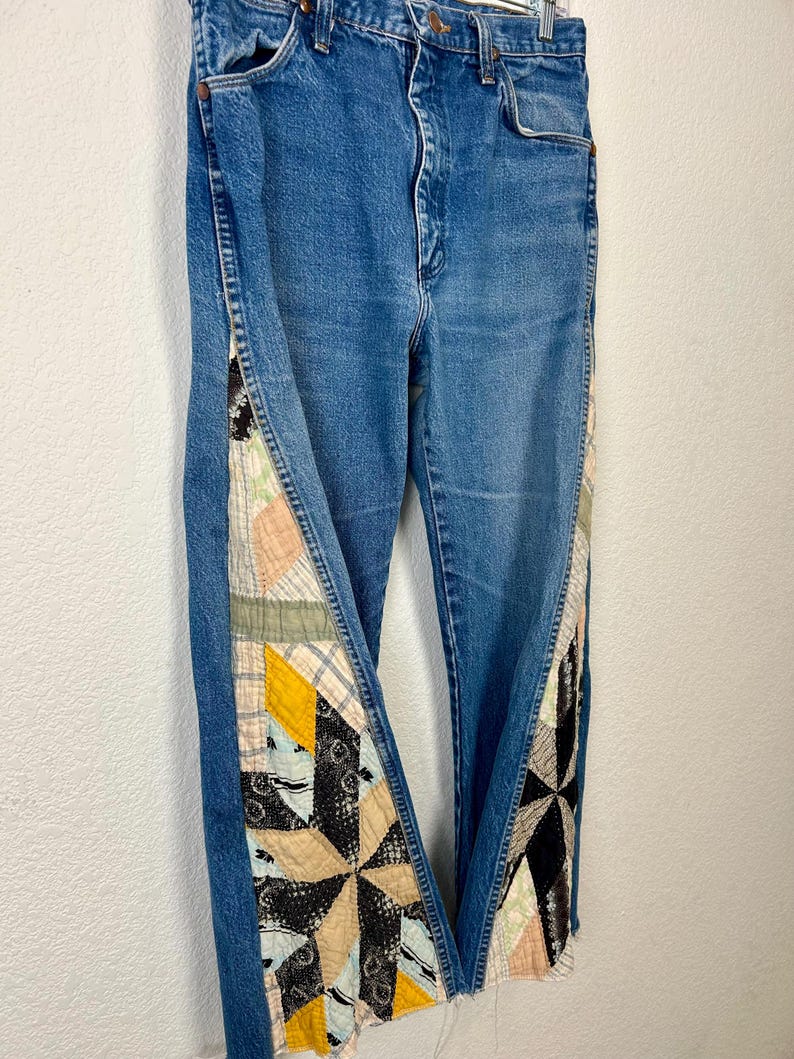 Op de afbeelding: Blauwe spijkerbroek met patchwork quilt details aan de zijkanten. Het patchwork heeft verschillende kleuren en patronen, waaronder beige, geel, zwart en roze. De jeans hebben een klassiek ontwerp met vijf zakken en een knoop- en ritssluiting.