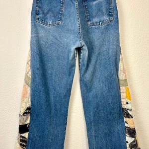 Op de afbeelding: Blauwe Wrangler-jeans met patchwork stoffen accenten op de buitenste naden. Het patchwork heeft een verscheidenheid aan patronen en kleuren, waaronder zwart, wit, geel en perzik. De jeans hebben een klassiek ontwerp met vijf zakken.