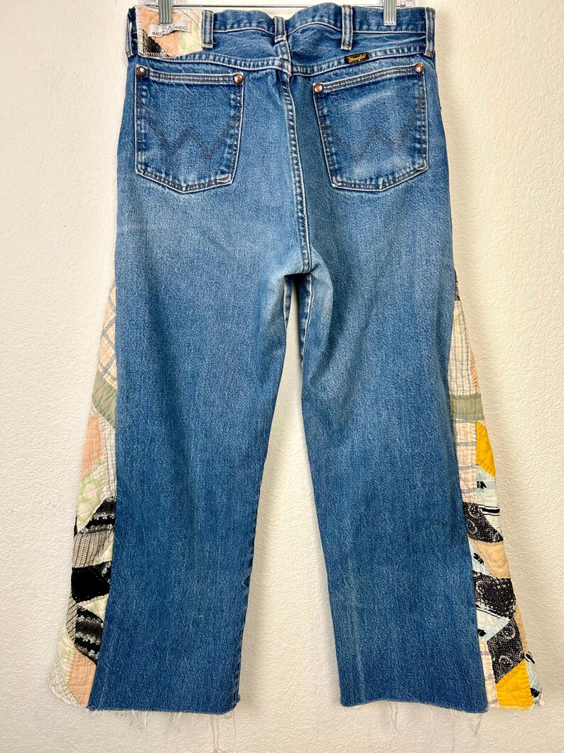 Op de afbeelding: Blauwe Wrangler-jeans met patchwork stoffen accenten op de buitenste naden. Het patchwork heeft een verscheidenheid aan patronen en kleuren, waaronder zwart, wit, geel en perzik. De jeans hebben een klassiek ontwerp met vijf zakken.