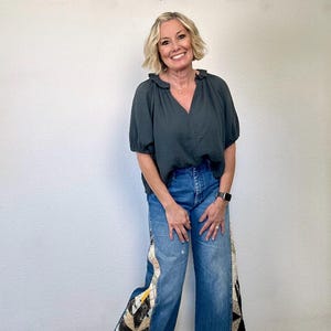Op de afbeelding: Een vrouw draagt een donkergroene blouse en een blauwe jeans met patchwork details. De jeans heeft een wijde pijp en een hoge taille. De patchwork heeft een verscheidenheid aan kleuren en patronen. De blouse heeft een V-hals en korte mouwen.