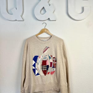 Könnte beinhalten: Beiges Sweatshirt mit einem Kürbis-Design aus Patchwork. Der Kürbis besteht aus verschiedenen Stoffmustern in Rot, Blau, Weiß und Schwarz. Das Sweatshirt hängt an einem Holzbügel vor einer weißen Wand.