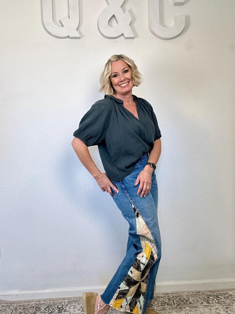 Op de afbeelding: Een persoon draagt een donkergroene top met korte mouwen en een blauwe jeans met patchwork details. De jeans heeft een gerafelde zoom en een decoratief paneel aan de zijkant met geometrische patronen. De achtergrond is een effen witte muur.