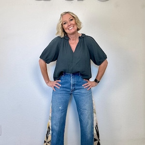 Op de afbeelding: Een vrouw draagt een donkergroene blouse en een blauwe spijkerbroek. De jeans heeft patchwork stoffen panelen aan de zijkanten. De achtergrond toont de letters "Q & C" in wit.