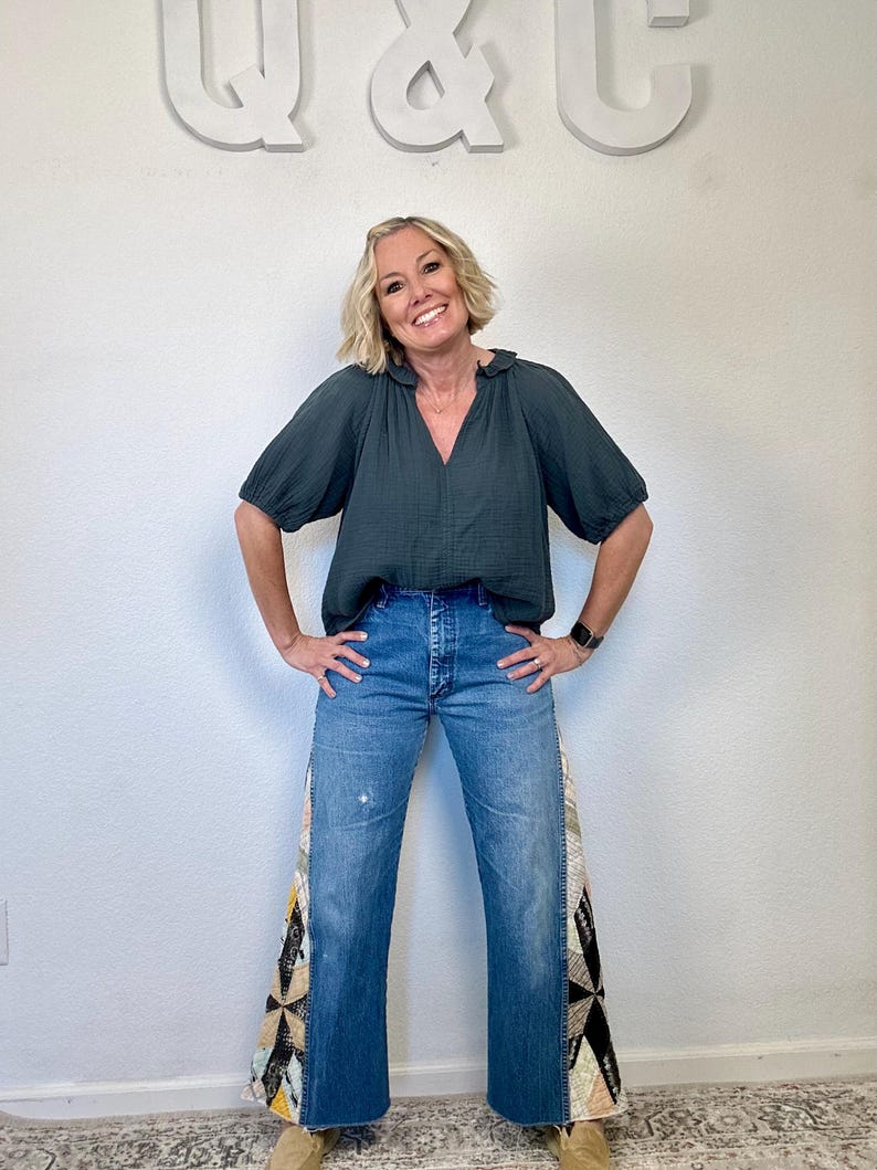 Op de afbeelding: Een vrouw draagt een donkergroene blouse en een blauwe spijkerbroek. De jeans heeft patchwork stoffen panelen aan de zijkanten. De achtergrond toont de letters "Q & C" in wit.