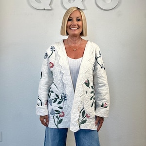 May include: A white quilted jacket featuring an open front, long sleeves, and a floral pattern with green leaves, blue berries, and pink flowers. The jacket is worn over a white ribbed tank top and light blue denim jeans.