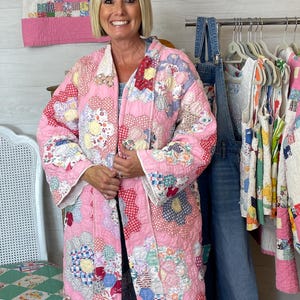 May include: A pink quilted coat with a floral and geometric patchwork design. The coat features a scalloped edge and is worn over blue denim overalls. A matching patchwork tank top hangs on a wooden hanger in the background. The coat is a unique, handmade garment.