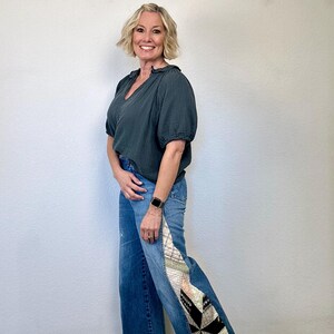 Op de afbeelding: Een vrouw draagt een donkergroene V-hals blouse en een blauwe jeans met een patchwork ontwerp aan de zijkant. De jeans hebben een wijde pijp en een rauwe zoom. De achtergrond is een witte muur met de letters "Q & Q" in wit.