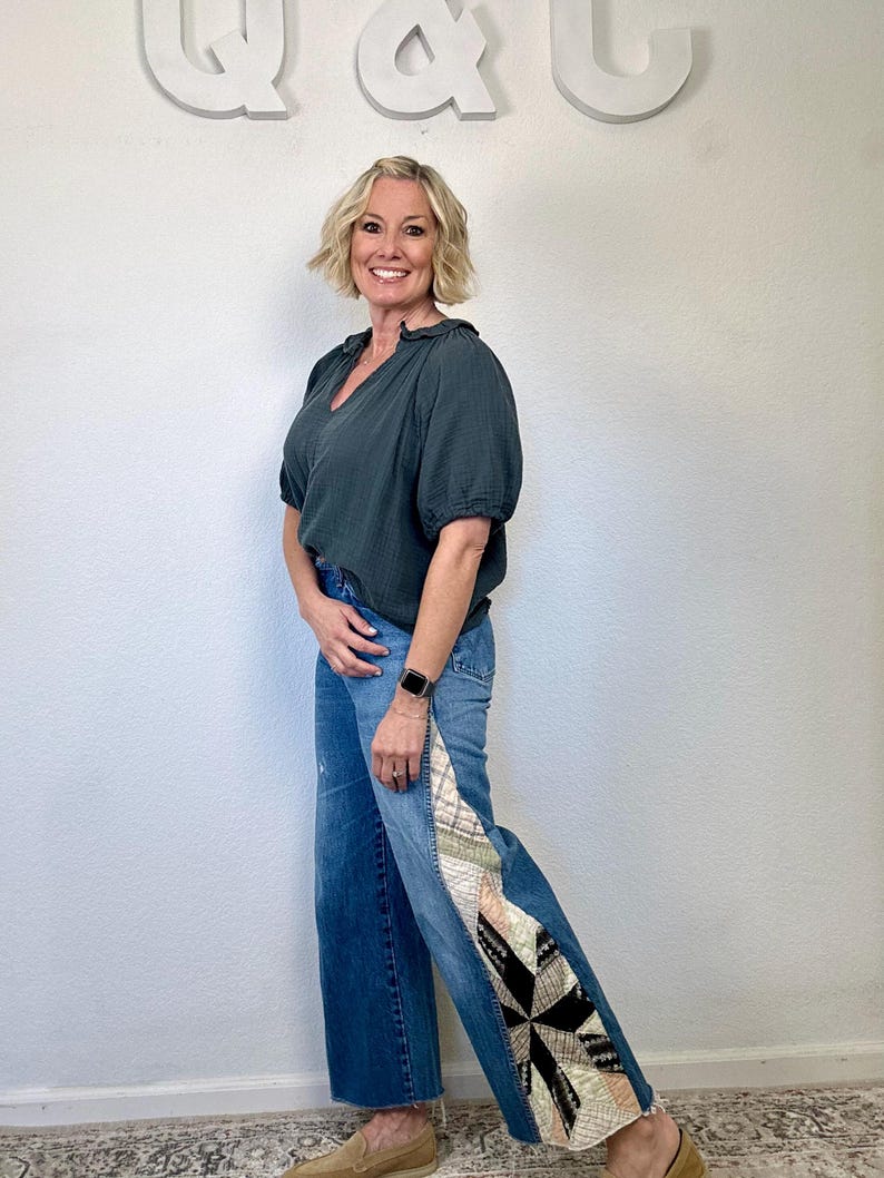 Op de afbeelding: Een vrouw draagt een donkergroene V-hals blouse en een blauwe jeans met een patchwork ontwerp aan de zijkant. De jeans hebben een wijde pijp en een rauwe zoom. De achtergrond is een witte muur met de letters "Q & Q" in wit.