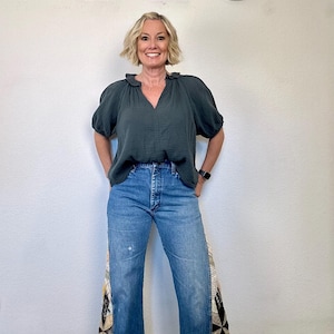 Op de afbeelding: Een vrouw draagt een donkergroene blouse met een gerimpelde halslijn en een blauwe jeans. De jeans hebben een patchwork ontwerp op de buitenste naden. De achtergrond is een witte muur met de letters "Q & C" in wit.