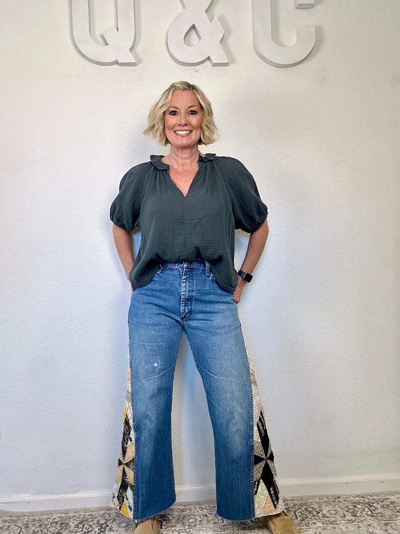 Op de afbeelding: Een vrouw draagt een donkergroene blouse met een gerimpelde halslijn en een blauwe jeans. De jeans hebben een patchwork ontwerp op de buitenste naden. De achtergrond is een witte muur met de letters "Q & C" in wit.
