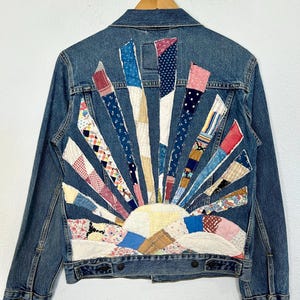 May include: A blue denim jacket with a patchwork quilt design on the back. The quilt design is a sun with rays and a cloud. The quilt is made of various fabrics in shades of blue, red, white, and yellow.