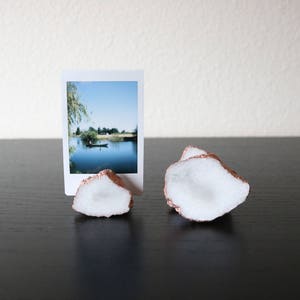 May include: Two white and copper-colored photo holders shaped like irregular rocks. The holders are on a dark surface and a photo of a lake with a boat is in the holder on the left.