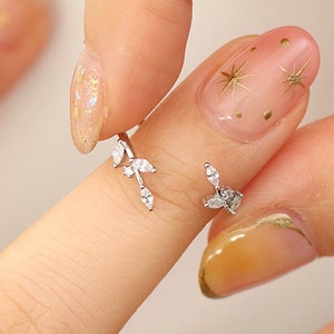 May include: A silver ring with a delicate leaf design featuring small clear stones. The ring is worn on a finger with a light pink nail polish and gold glitter accents.