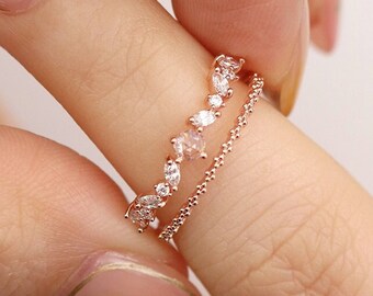 May include: Two delicate rose gold rings with clear cubic zirconia stones. The top ring features a curved row of stones, while the bottom ring is a simple chain.