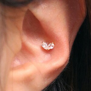 Puede incluir: Primer plano de la oreja de una persona con un pequeño pendiente de corazón de oro con piedras transparentes. El pendiente está en el cartílago superior de la oreja.