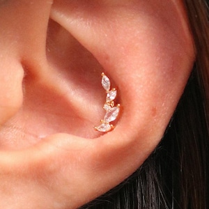 Puede incluir: Un piercing dorado con un diseño curvo. El piercing presenta varias piedras preciosas claras, de talla marquesa, dispuestas en forma de hoja, añadiendo un toque de brillo. La joya está colocada sobre la piel de la oreja.