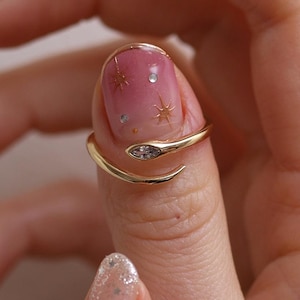 May include: A gold snake ring with a clear gemstone at the head. The ring is wrapped around a finger. The background is a hand with pink and white nail polish. The nail polish has star and gem designs.