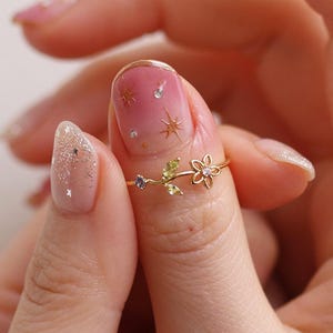May include: A delicate gold ring featuring a floral design with a small flower and leaves. The ring is adorned with small gemstones and is worn on a finger. The ring's design is intricate and elegant.