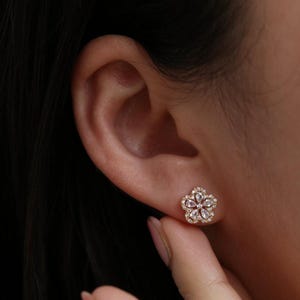 May include: A close-up of a flower-shaped stud earring. The earring is gold-toned with a cluster of small, clear stones at the center and a halo of smaller, clear stones around the petals. The earring is worn on an ear.