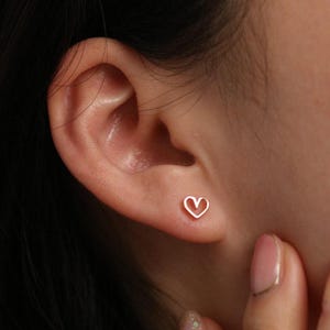 May include: A close-up of a person's ear with a small, heart-shaped earring. The earring is a simple outline of a heart, and it appears to be made of a gold-colored metal. The skin tone is light, and the earring is the focal point.