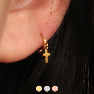 May include: Gold hoop earrings with a small cross charm hanging from the bottom. The earrings are worn in the ear of a person with dark hair.