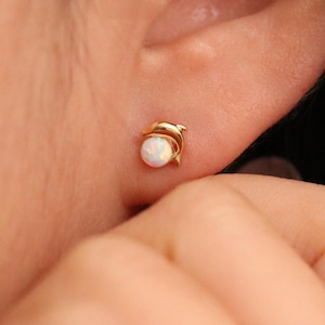 May include: A pair of gold dolphin earrings with a white opal gemstone. The earrings are small and delicate, perfect for everyday wear.