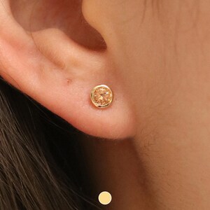 May include: A gold stud earring with a round, light orange gemstone. The earring is set in a gold bezel and is worn on an ear. The skin tone is light. The earring is a small piece of jewellery.