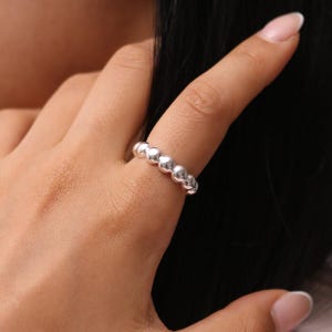 Puede incluir: Un anillo de plata con un diseño de cuentas. El anillo está formado por una serie de pequeñas cuentas redondas que están conectadas entre sí para formar una banda continua.
