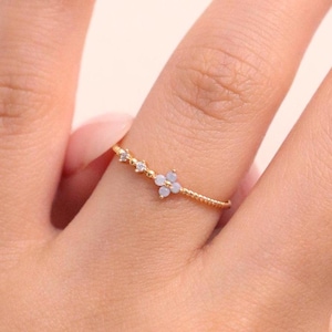 May include: A delicate gold ring with a floral design, featuring small, light blue gemstones and clear crystals. The ring is worn on a finger, highlighting its intricate details and elegant style. The band has a textured design.