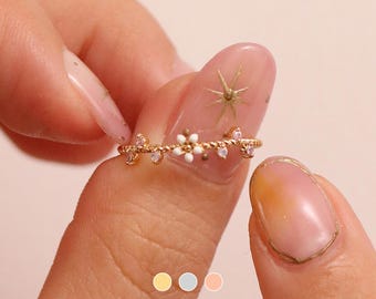 May include: A gold ring with a delicate vine design and small white flowers. The ring is worn on a finger with a light pink nail polish and a gold star design.